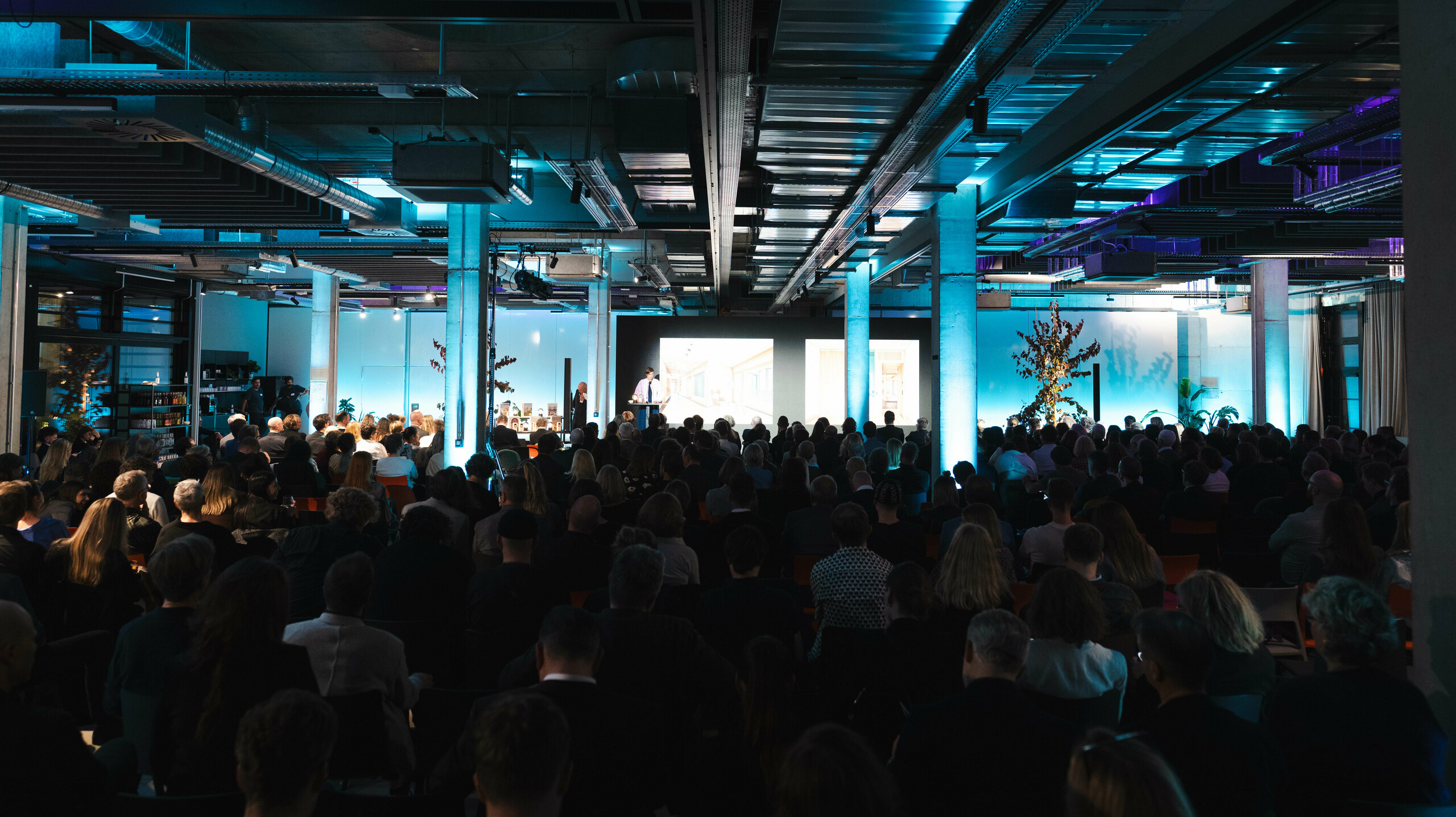 Nastrojowa scena z ceremonii wręczenia nagród „Budynek mieszkalny roku 2025”: widok na wypełnioną po brzegi salę w nowoczesnej hali przemysłowej z efektownym niebieskim oświetleniem. Na pierwszym planie znajduje się scena z dużym ekranem, na którym wyświetlane są prezentacje. Architektura pomieszczenia z odsłoniętymi stalowymi belkami i betonowymi filarami podkreśla innowacyjny charakter wydarzenia, podczas którego świętowane są zrównoważone rozwiązania, takie jak moduł solarny PREFALZ.