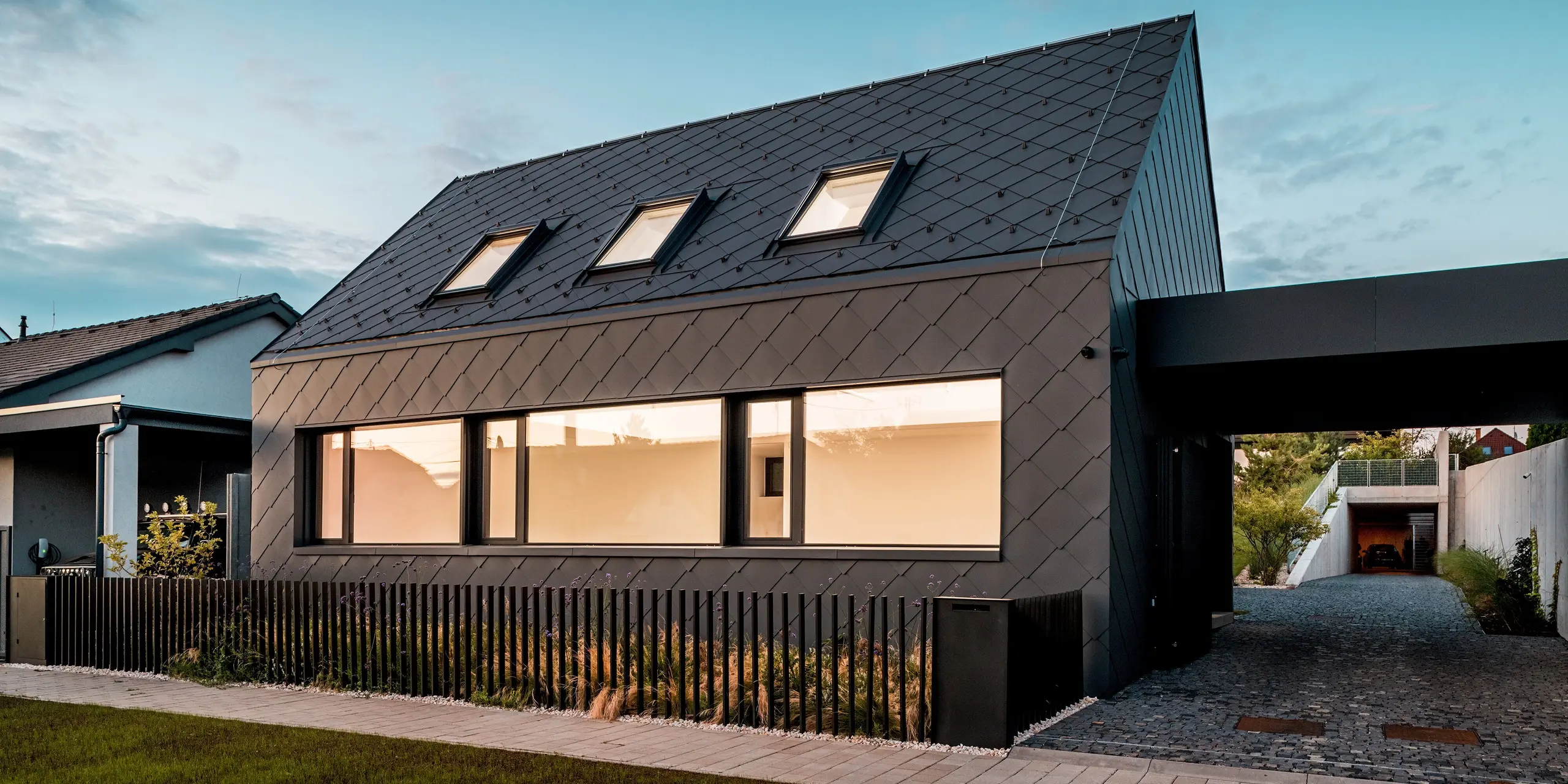 Das Bild zeigt das moderne Einfamilienhaus in Trnava‑Modranka in der Abenddämmerung, komplett verkleidet mit PREFA Dach- und Wandrauten 44 × 44 in P.10 Dunkelgrau. Die beleuchteten Fenster erzeugen einen warmen Kontrast zur dunklen, gleichmäßig strukturierten Aluminiumhülle. Die präzise verlegte Rautenoptik betont die klaren Linien des Baukörpers und unterstreicht die hochwertige, robuste Materialität. Die langlebigen PREFA Aluminiumprodukte bieten optimalen Witterungsschutz und verleihen dem Gebäude ein markantes, zeitloses Erscheinungsbild.