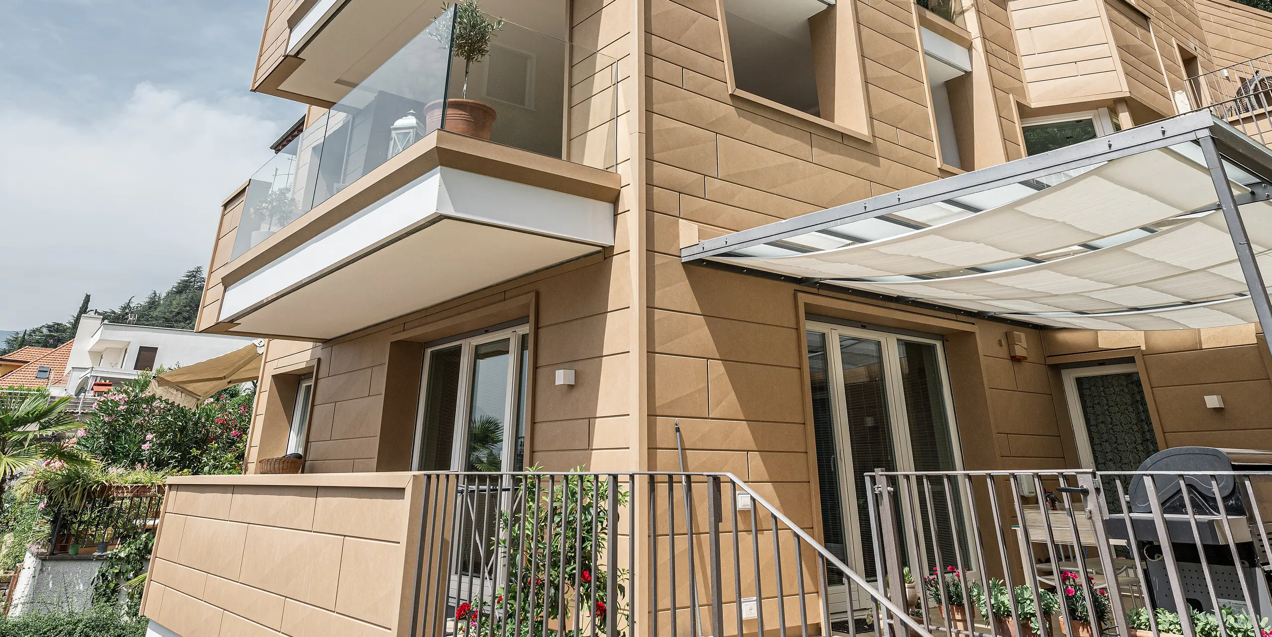 Blick aufwärts an der Fassade der Villa Laura, kunstvoll gestaltet mit PREFA Siding.X in Sandbraun, die moderne Architektur und Funktionalität in einer langlebigen Aluminiumhülle vereint. Überhängende Balkone und große Fensterflächen öffnen das Gebäude zum Himmel, während die feine Textur der Fassadenpaneele und deren natürliche Farbgebung das Gebäude optisch erden und in die mediterrane Landschaft Südtirols einbetten.