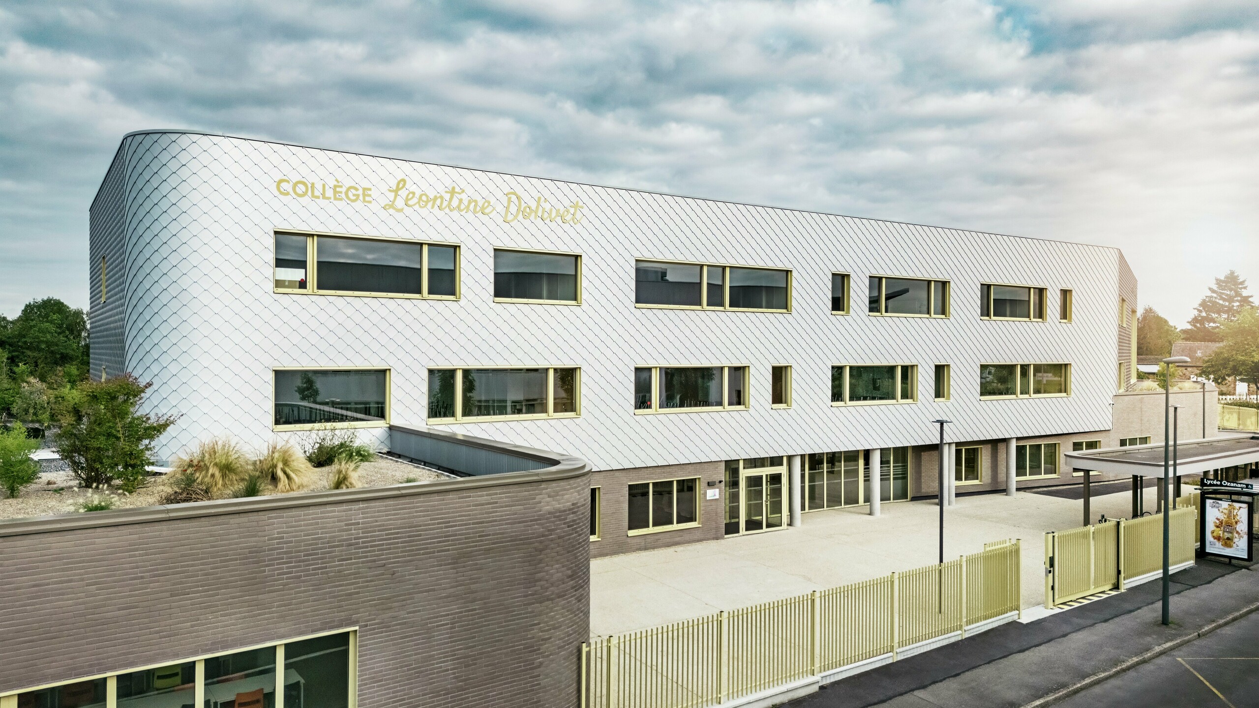 Die Vorderansicht des Collège Léontine Dolivet in Cesson-Sévigné zeigt eine moderne Aluminiumfassade aus PREFA-Wandelementen in Silbermetallic. Die variable Oberfläche verleiht dem Gebäude Leichtigkeit, während klare Linien und große Fenster für Transparenz sorgen. Gelbe Fensterrahmen setzen farbliche Akzente. Im Vordergrund sind der helle Eingangsbereich, der Zaun und die angrenzende Straße zu sehen. Die langlebige PREFA-Fassadenlösung vereint Design, Witterungsbeständigkeit und Nachhaltigkeit – ein architektonisch ansprechendes Gebäude mit einer eleganten Formensprache.