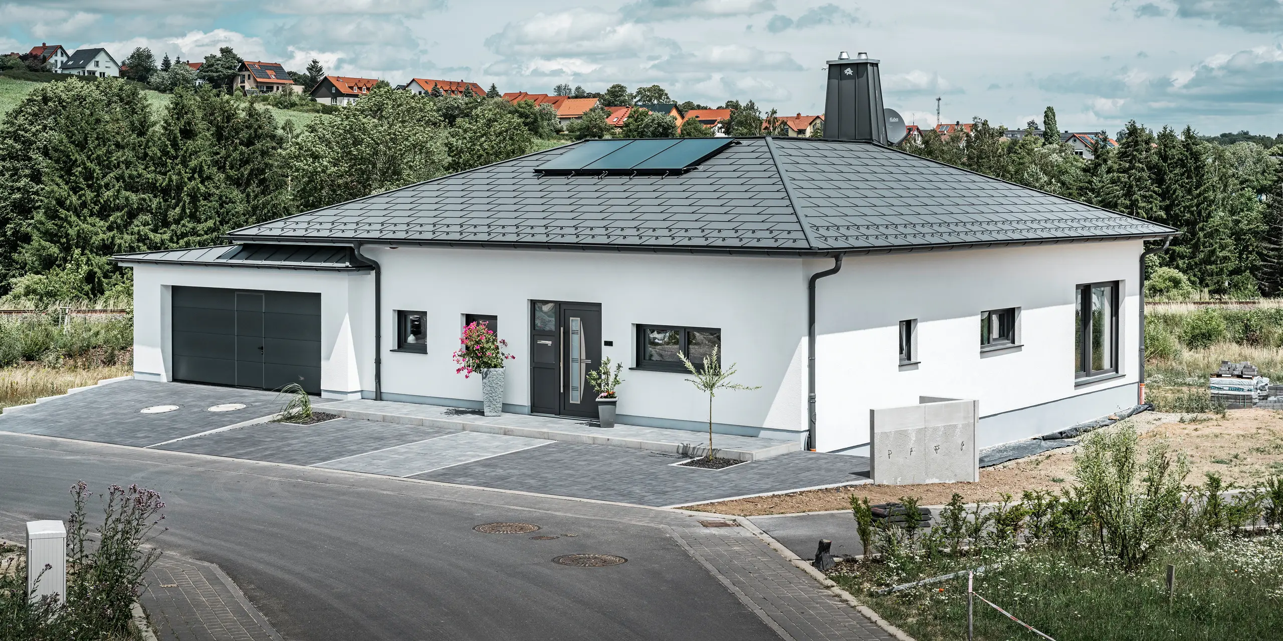 Elegancki dom jednorodzinny w Ilmenau w Turyngii, z wysokiej jakości dachem PREFA w kolorze P.10 grafitowym, wyposażonym w wytrzymałą dachówkę R.16. Garaż harmonizuje pod względem koloru i stylu dzięki zastosowaniu PREFALZ. Atrakcją nieruchomości jest funkcjonalnie zaprojektowane atrium oraz komin ozdobiony symbolem PREFA Stier, które podkreślają nowoczesny design i innowacyjną technologię dachową PREFA. Panele słoneczne idealnie wpisują się w zrównoważoną koncepcję domu. Przemyślaną koncepcję bungalowu dopełnia system odprowadzania wody z dachu PREFA, również w kolorze grafitowym.