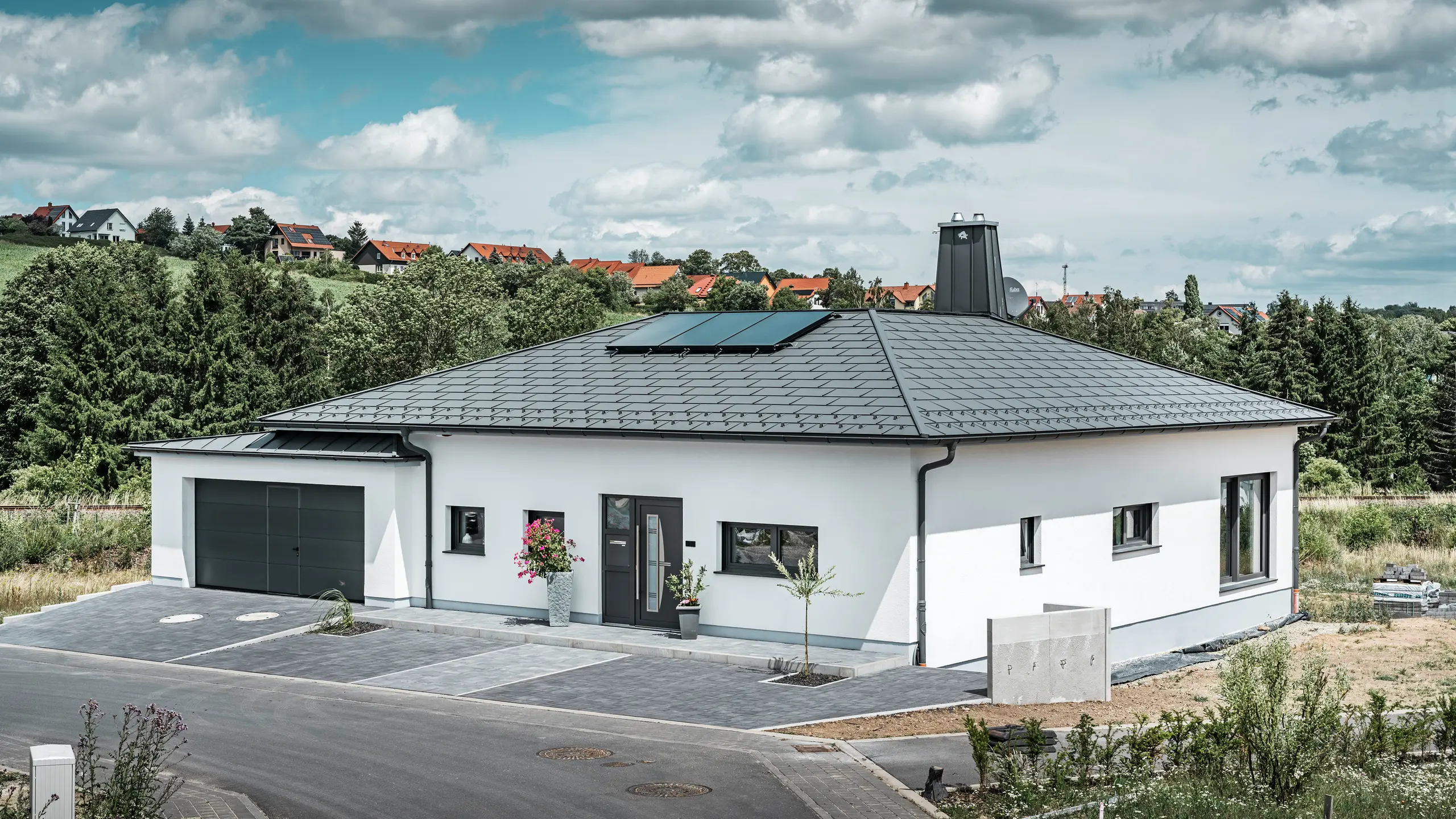Elegancki dom jednorodzinny w Ilmenau w Turyngii, z wysokiej jakości dachem PREFA w kolorze P.10 grafitowym, wyposażonym w wytrzymałą dachówkę R.16. Garaż harmonizuje pod względem koloru i stylu dzięki zastosowaniu PREFALZ. Atrakcją nieruchomości jest funkcjonalnie zaprojektowane atrium oraz komin ozdobiony symbolem PREFA Stier, które podkreślają nowoczesny design i innowacyjną technologię dachową PREFA. Panele słoneczne idealnie wpisują się w zrównoważoną koncepcję domu. Przemyślaną koncepcję bungalowu dopełnia system odprowadzania wody z dachu PREFA, również w kolorze grafitowym.