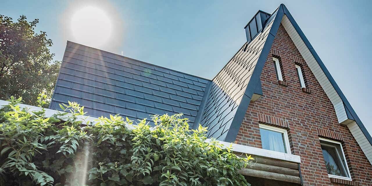 Dach dwuspadowy pokryty gładką dachówką aluminiową PREFA R.16 w kolorze antracytowym. Elewacja o rustykalnym charakterze jest wykonana z klinkieru, za domem wschodzi właśnie słońce.