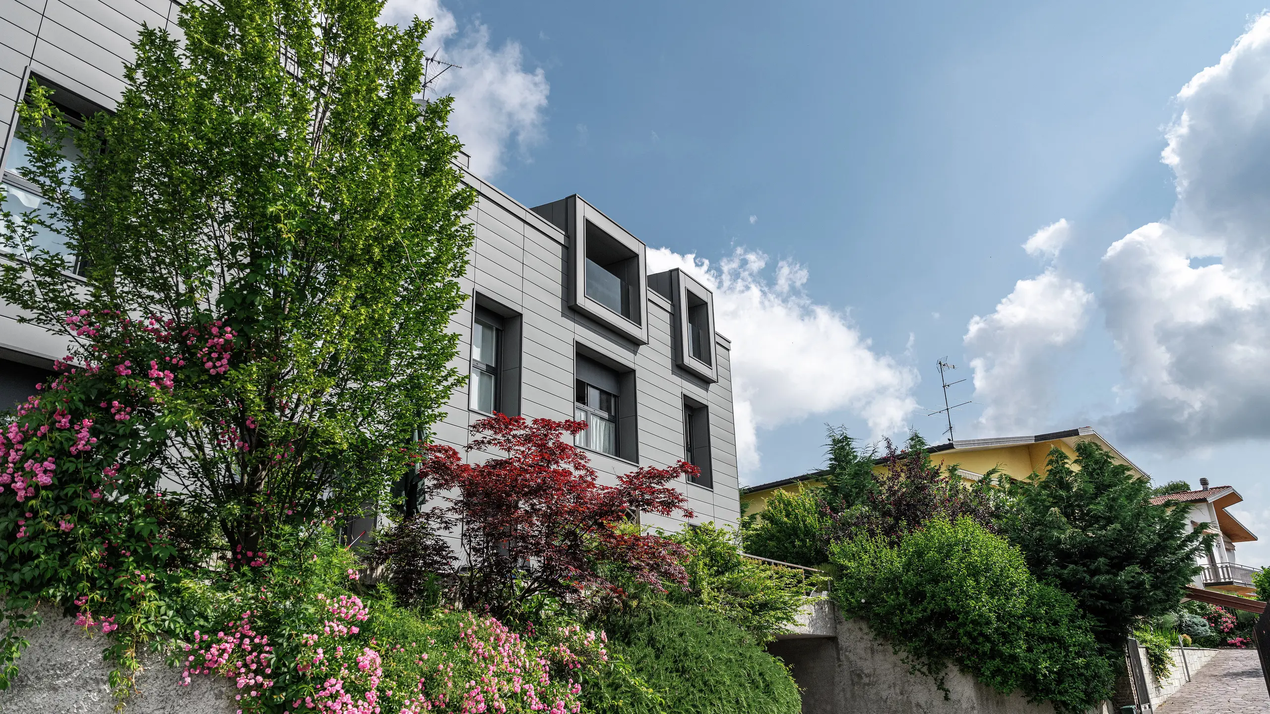 Nowoczesny kompleks mieszkaniowy w Faloppio we Włoszech, z elegancką elewacją wykonaną z okładzin PREFA. Wystające okna stanowią wyjątkowy element architektoniczny, a otaczające je rośliny i kwiaty podkreślają naturalne piękno obiektu. Trwała okładzina aluminiowa PREFA stanowi estetyczne i funkcjonalne rozwiązanie na każdą pogodę.