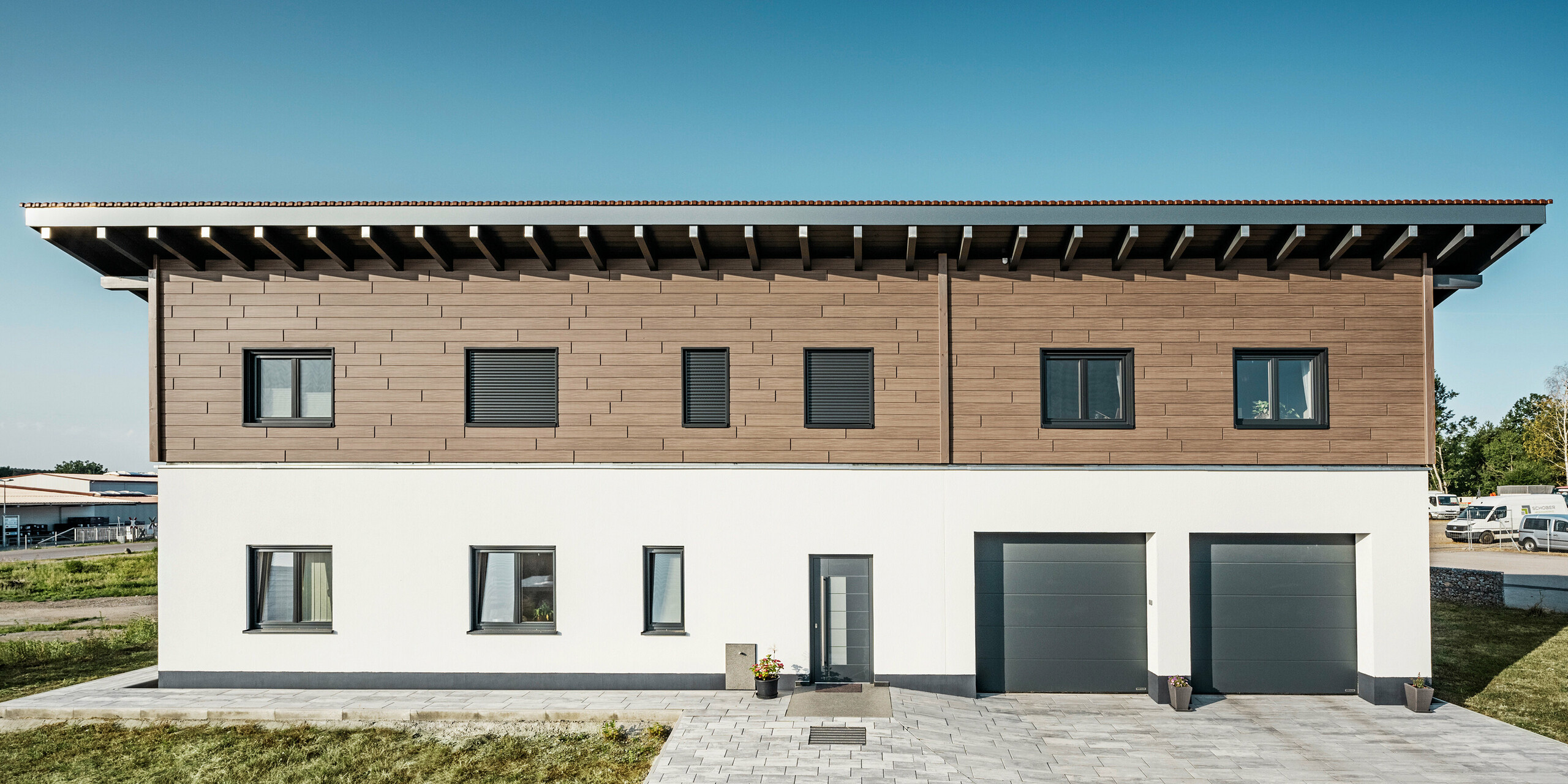 Blick von vorne auf ein Einfamilienhaus in Bruck mit einer Aluminiumfassade in einer Holzoptik am Obergeschoss. Das obere Stockwerk wurde mit PREFA Sidings in der Farbe Walnuss Braun verkleidet.