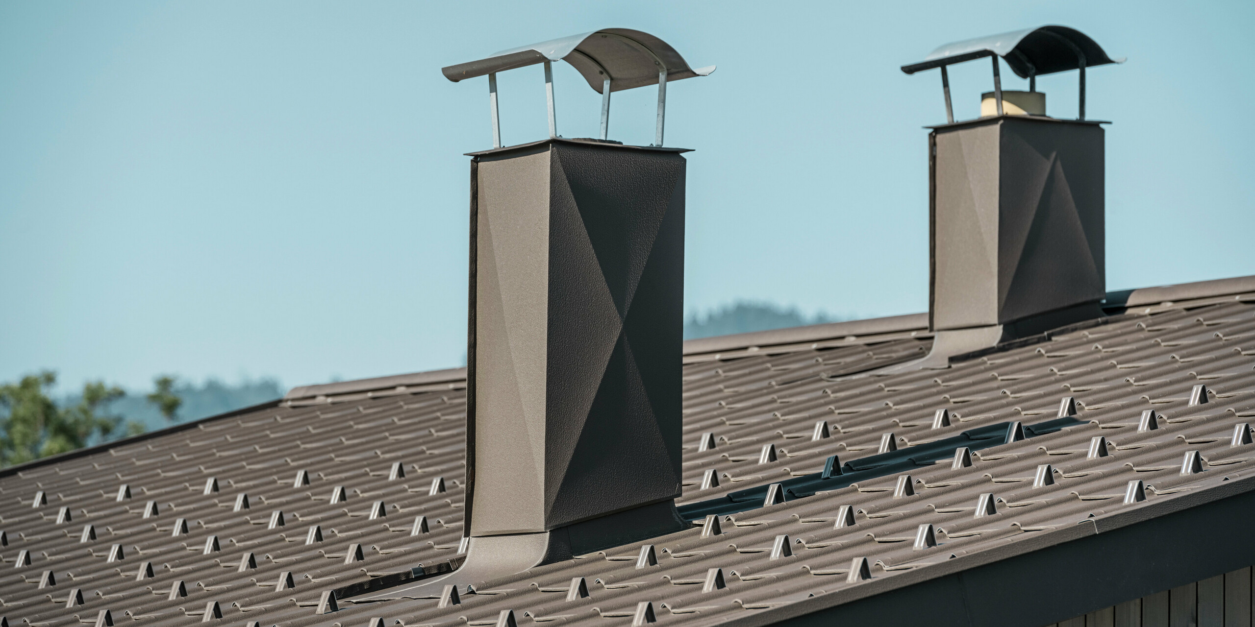 Detailaufnahme des Daches eines traditionell österreichischen Einfamilienhauses aus Holz, welches mit PREFA Dachplatten in P.10 Braun eingedeckt wurde. Auf dem Bild stechen zwei moderne Kaminverkleidungen hervor, welche von PREFA Aluminiumprodukten in P.10 Braun ummantelt sind. Die Ästhetik des Daches harmoniert hervorragend mit der Holzfassade und schafft ein einheitliches, natürlich wirkendes Erscheinungsbild, das die langlebigen und wartungsarmen Eigenschaften von PREFA Produkten unterstreicht.