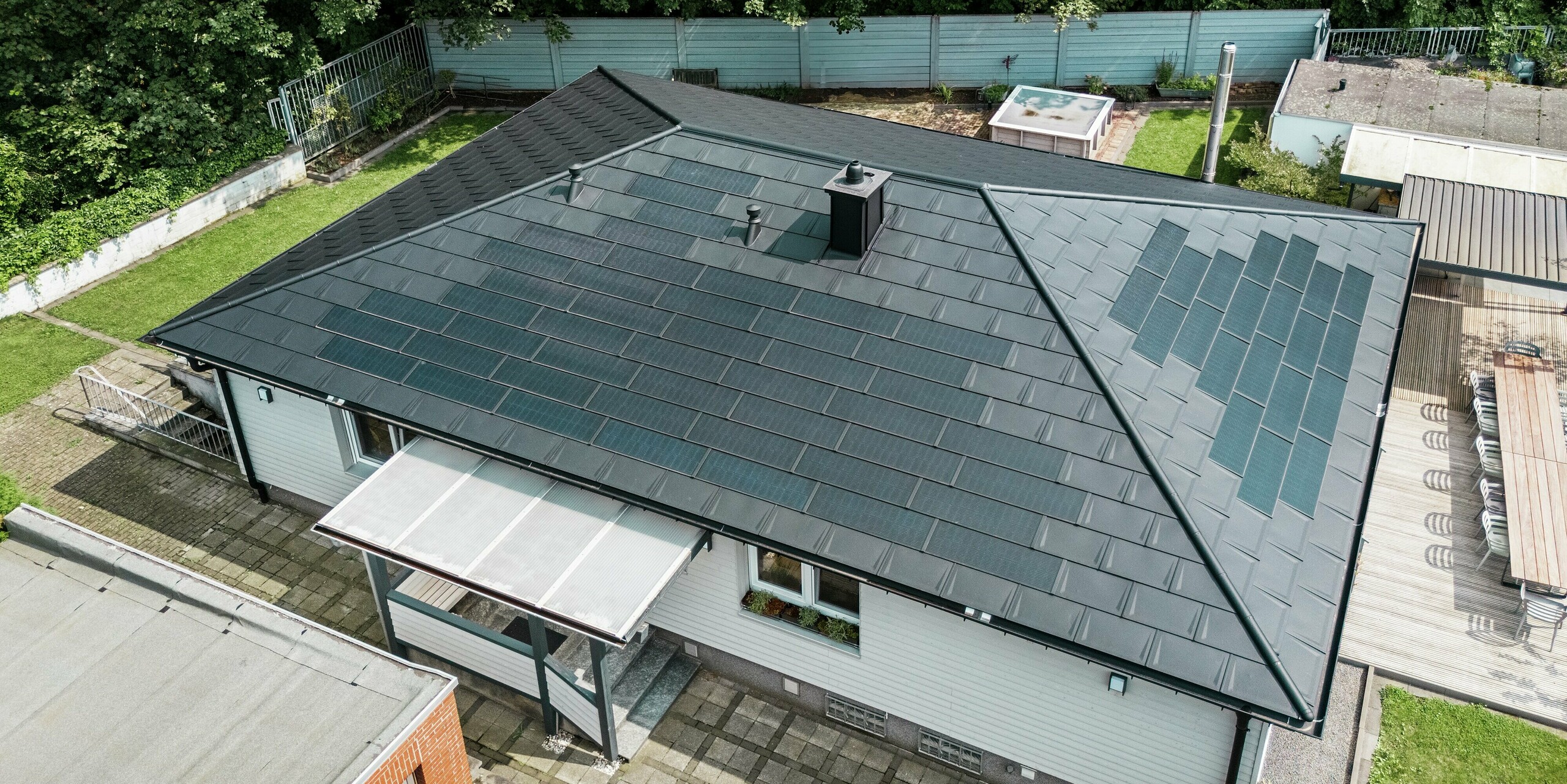 Moderner Bungalow in Rommerskirchen aus der Drohnenperspektive mit Blick auf das nachhaltige PREFA Aluminiumdach. Die innovativen PREFA Solardachplatten erzeugen Strom aus Sonnenenergie. Rund um die Aluminiumdachplatten mit integrierten Photovoltaikmodulen sorgen die robusten PREFA Dachplatten R.16 für maximalen Schutz. Die Dachentwässerung mit Saumrinne und Ablaufrohr in P.10 schwarz bietet Funktionalität und Ästhetik für ein harmonisches Gesamtbild.