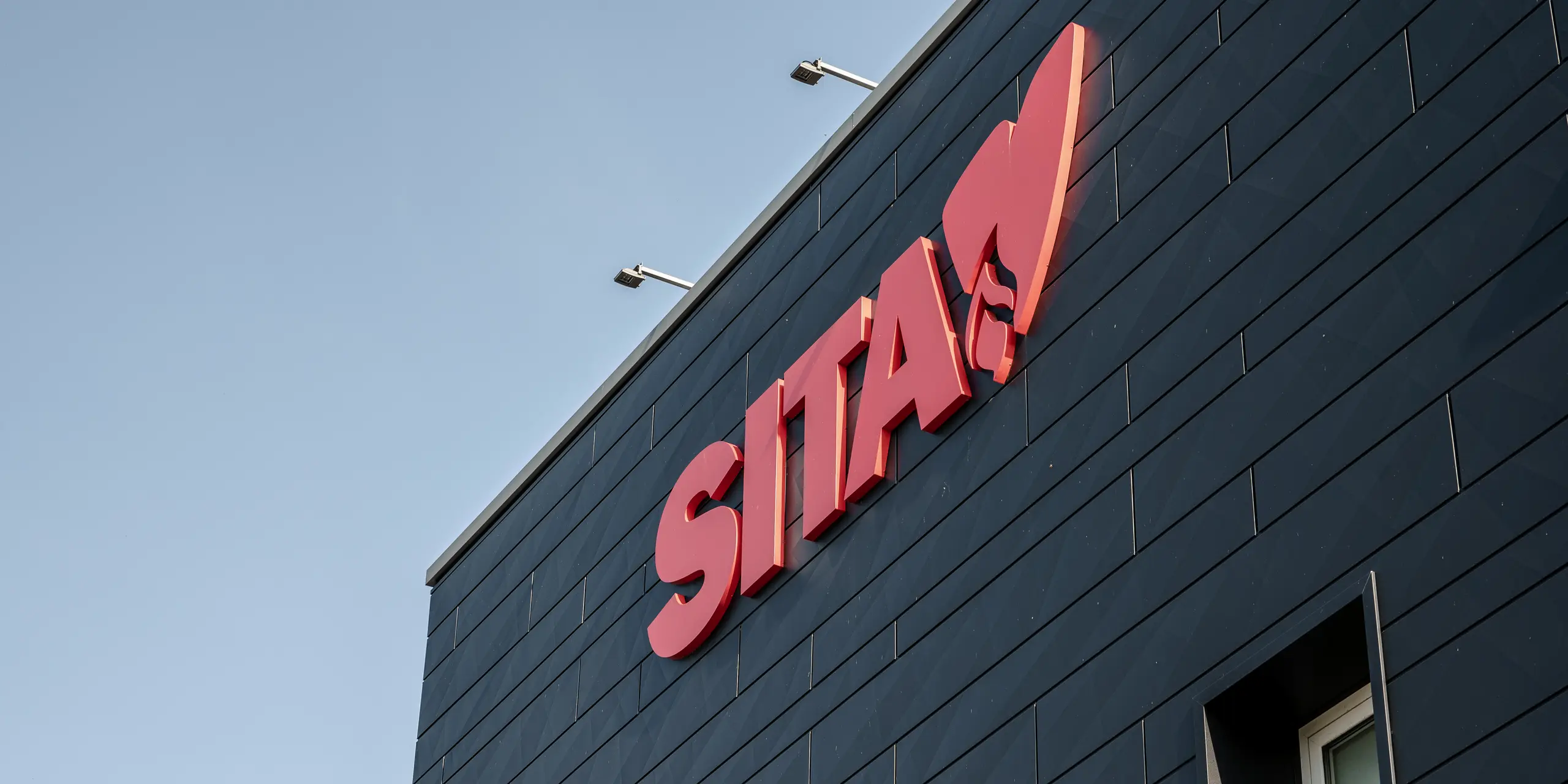 Nahaufnahme des SITA-Firmenschildes in einem auffälligen Rot, kontrastreich hervorgehoben gegen die strukturierte dunkelgraue PREFA Siding.X Fassade in P.10 Schwarz. Das Bild zeigt die präzise Verarbeitung und die charakteristische Oberflächenstruktur der Aluminiumpaneele, die für eine moderne, ästhetische Fassadengestaltung sorgen und die Corporate Identity von SITA in Porcellengo, Italien, unterstreichen. Die Außenbeschilderung setzt zusätzliche Akzente und betont die qualitativ hochwertige und innovative Fassadenlösung von PREFA, die sowohl visuell ansprechend als auch funktional ist.