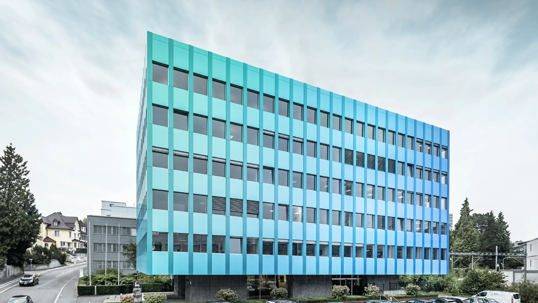 Widok boczny budynku Wielandhaus w Aarau; elewacja pokryta jest płytą kompozytową PREFABOND z aluminium firmy PREFA, która dzięki specjalnej technologii powlekania mieni się niebiesko-zielonym blaskiem.