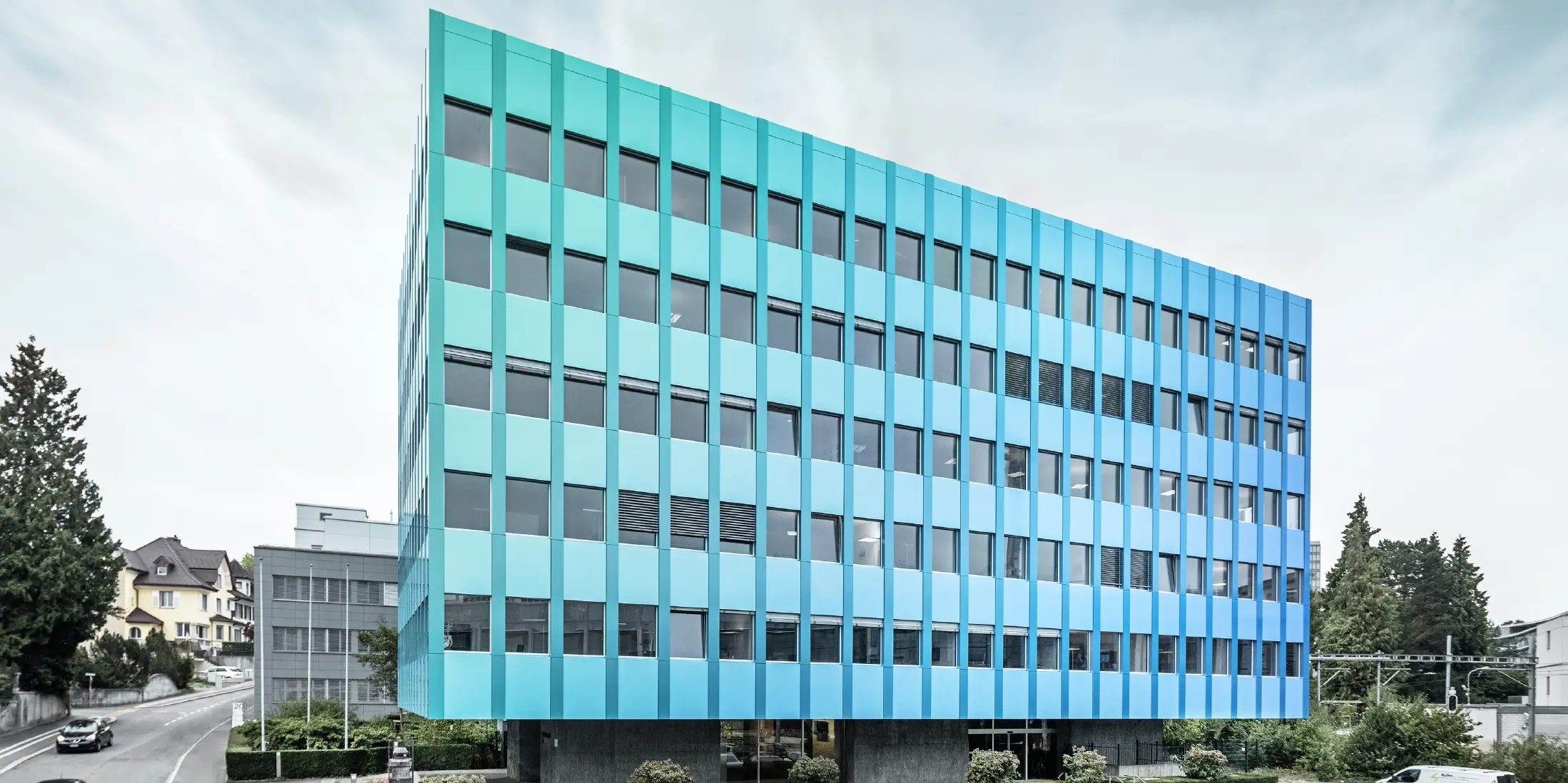 Widok boczny budynku Wielandhaus w Aarau; elewacja pokryta jest płytą kompozytową PREFABOND z aluminium firmy PREFA, która dzięki specjalnej technologii powlekania mieni się niebiesko-zielonym blaskiem.