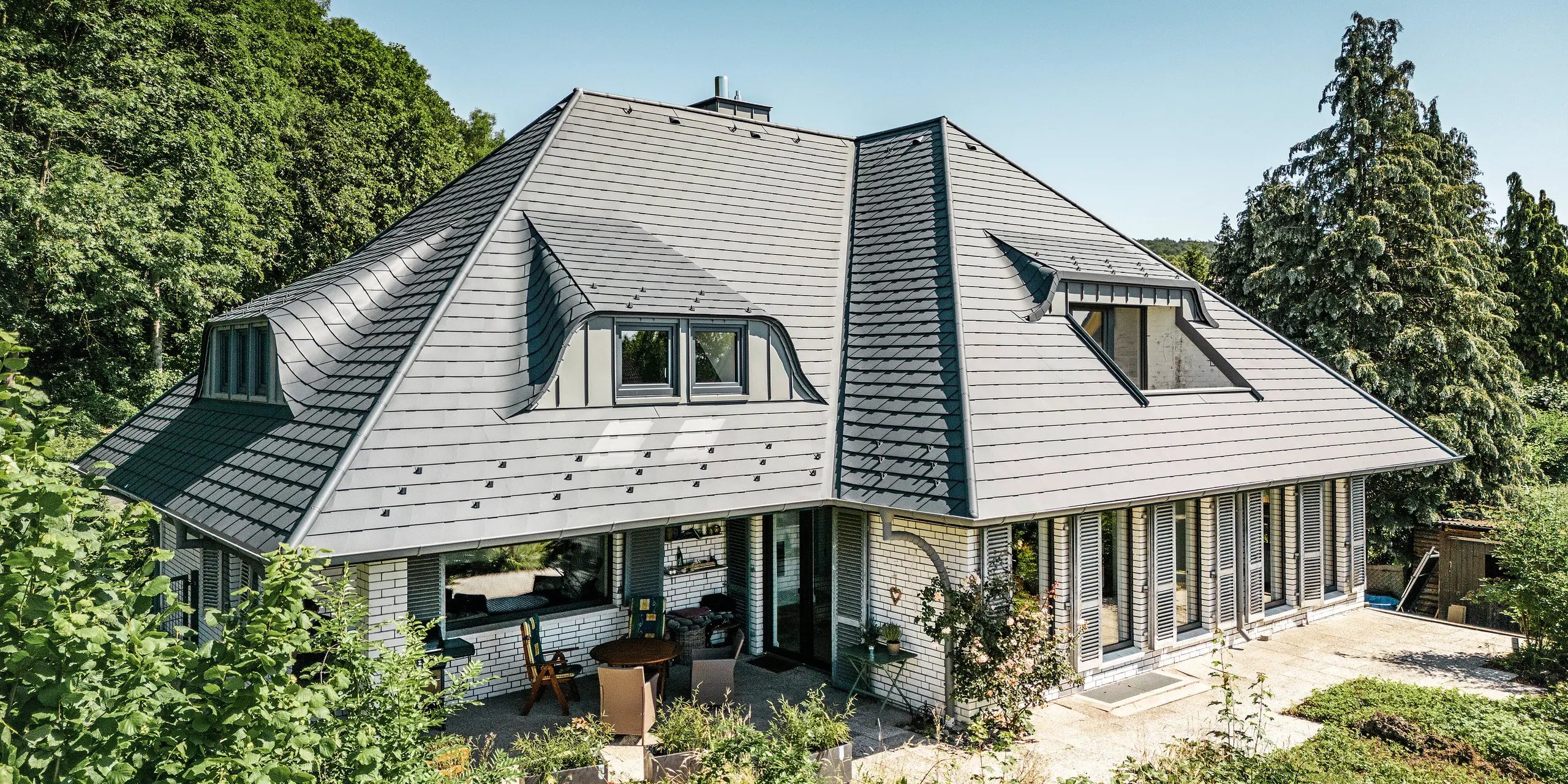 Nowoczesny dom jednorodzinny w Einbeck w Niemczech, pokryty gontem dachowym PREFA w kolorze P.10 antracyt. Trwały gont dachowy PREFA z aluminium znany jest ze swojej jakości i trwałości. Konstrukcja dachu budynku podkreśla indywidualną architekturę dzięki lukarnom, które zostały uwydatnione dzięki elastyczności gontu dachowego PREFA. Odwodnienie dachu w postaci rury spustowej PREFA lub rynny wiszącej również utrzymane jest w kolorze antracytowym. Wokół domu jednorodzinnego znajduje się obszerny i zadbany ogród oraz taras. Za domem rozciąga się las, który wraz z błękitnym niebem tworzy naturalną atmosferę.
