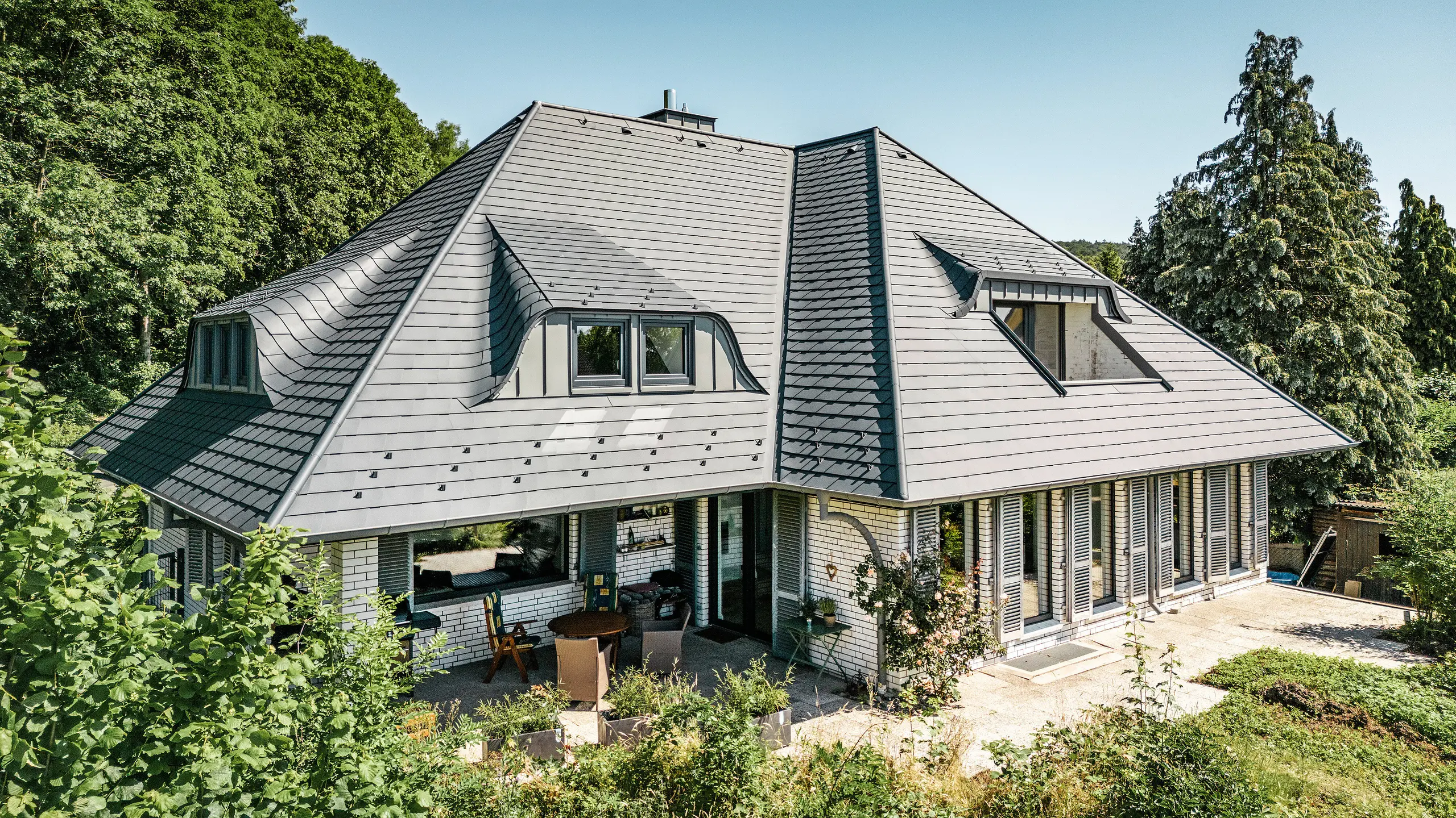 Nowoczesny dom jednorodzinny w Einbeck w Niemczech, pokryty gontem dachowym PREFA w kolorze P.10 antracyt. Trwały gont dachowy PREFA z aluminium znany jest ze swojej jakości i trwałości. Konstrukcja dachu budynku podkreśla indywidualną architekturę dzięki lukarnom, które zostały uwydatnione dzięki elastyczności gontu dachowego PREFA. Odwodnienie dachu w postaci rury spustowej PREFA lub rynny wiszącej również utrzymane jest w kolorze antracytowym. Wokół domu jednorodzinnego znajduje się obszerny i zadbany ogród oraz taras. Za domem rozciąga się las, który wraz z błękitnym niebem tworzy naturalną atmosferę.