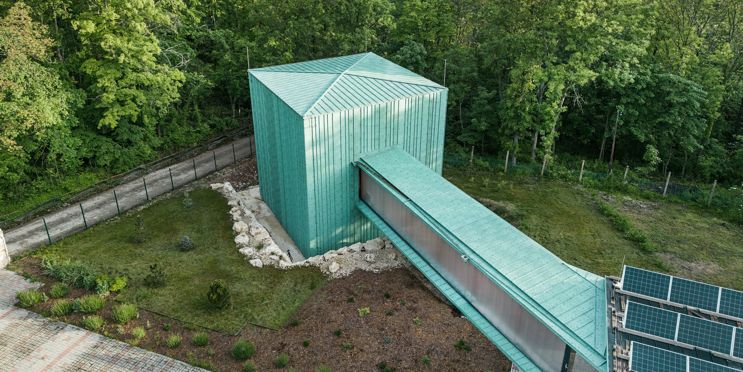 Das Observatorium Fertöboz aus der Vogelperspektive: ein architektonisch markantes Gebäude, dessen Dach und Fassade mit patinagrünem PREFALZ gestaltet sind. Das Dach- und Fassadensystem aus Aluminium zeigt ein einheitliches vertikales Muster, das die klaren Linien und die moderne Architektur des Bauwerks betont. Der Komplex ist von Bäumen umgeben und fügt sich harmonisch in die natürliche Umgebung ein, während die Photovoltaikanlage auf dem Boden die nachhaltige Ausrichtung der Einrichtung unterstreicht. Im Hintergrund erstreckt sich eine offene Landschaft, was den Kontrast zwischen der dichten Bewaldung und dem offenen Raum hervorhebt. Das Bauwerk steht als ein Beispiel für moderne, umweltbewusste Bauweise, die sowohl ästhetisch ansprechend als auch funktionell und langlebig ist.