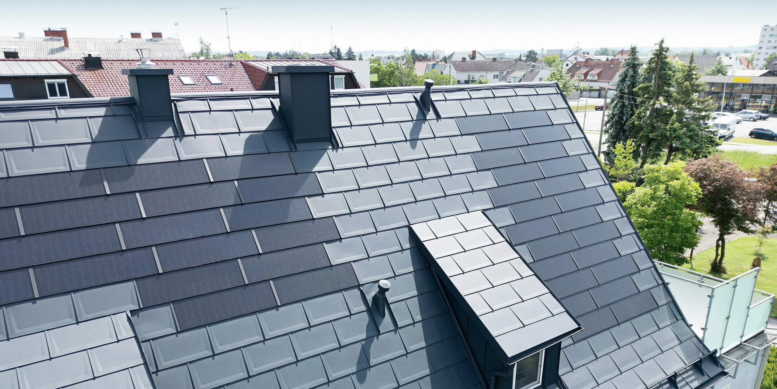 Vollständig saniertes Dach eines Einfamilienhauses in Leonding mit PREFA Solardachplatten in Anthrazit. Die Nahaufnahme zeigt das modernisierte Blech-Dach mit integrierten Solarmodulen von PREFA. Diese langlebige und witterungsbeständige Lösung bietet eine nachhaltige und energieeffiziente Möglichkeit zur Stromerzeugung. Das Bild verdeutlicht die ästhetische Integration der Solarmodule in die Dachfläche, die eine moderne und funktionale Architektur unterstützt. Ein zusätzlicher Eyecatcher der Dachlandschaft sind die langen Schleppgauben, verkleidet mit Dachplatten R.16.