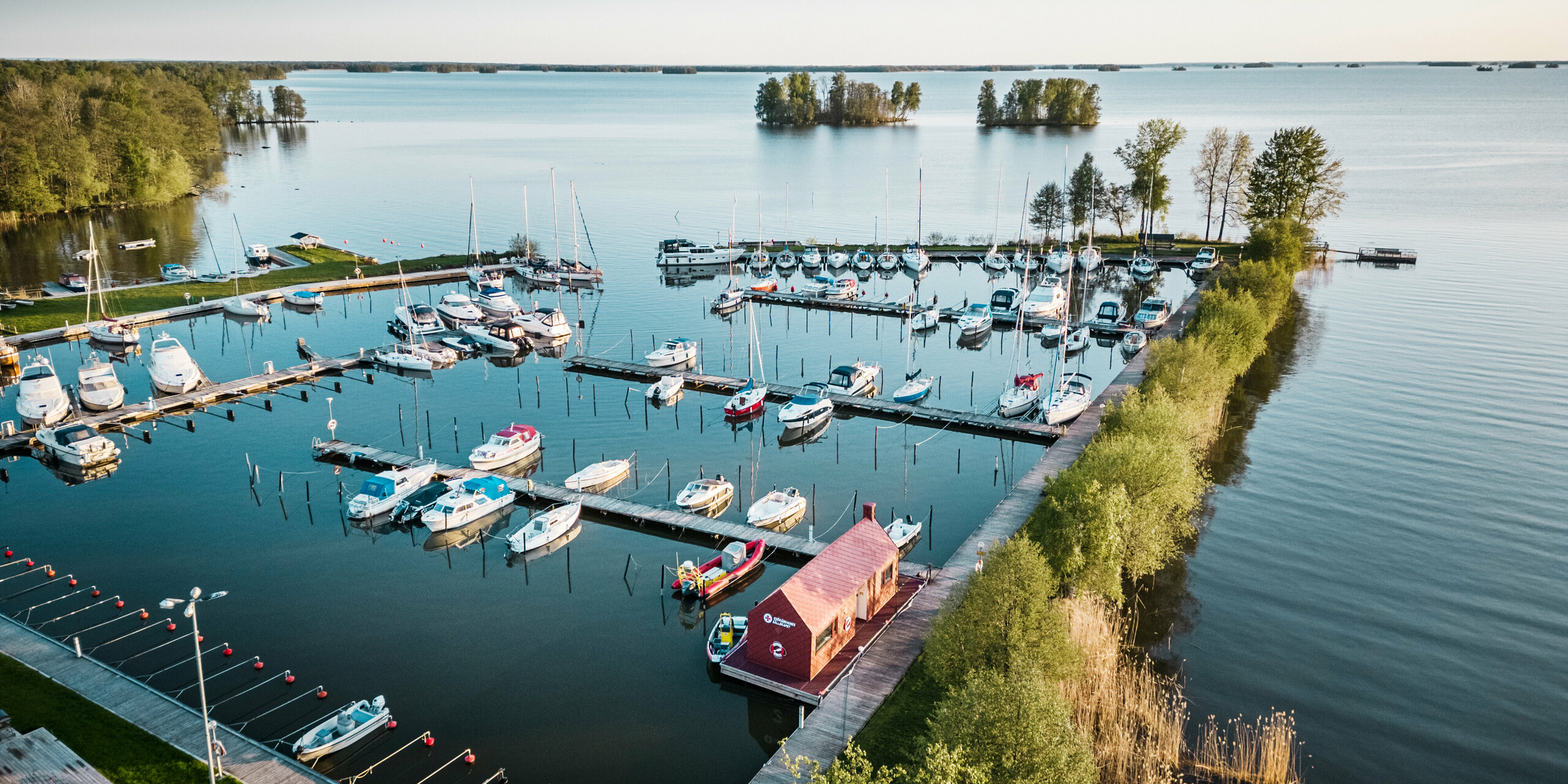 Luftbild des Seenotrettungshauses im Hafen am Hjälmaren See, Schweden, eingebettet zwischen Segelbooten und ruhigem, blauen Wasser. Das auffällige oxydrote Dach und die Fassade des Hauses aus PREFA Dach- und Wandrauten 29×29 fügen sich harmonisch in das maritime Umfeld ein und setzen zugleich einen starken visuellen Akzent. Die Rettungsstation dient als lebenswichtige Einrichtung für die Seenotretter und zeugt von der Verbindung zwischen Mensch und Natur sowie der Notwendigkeit, beides zu schützen.