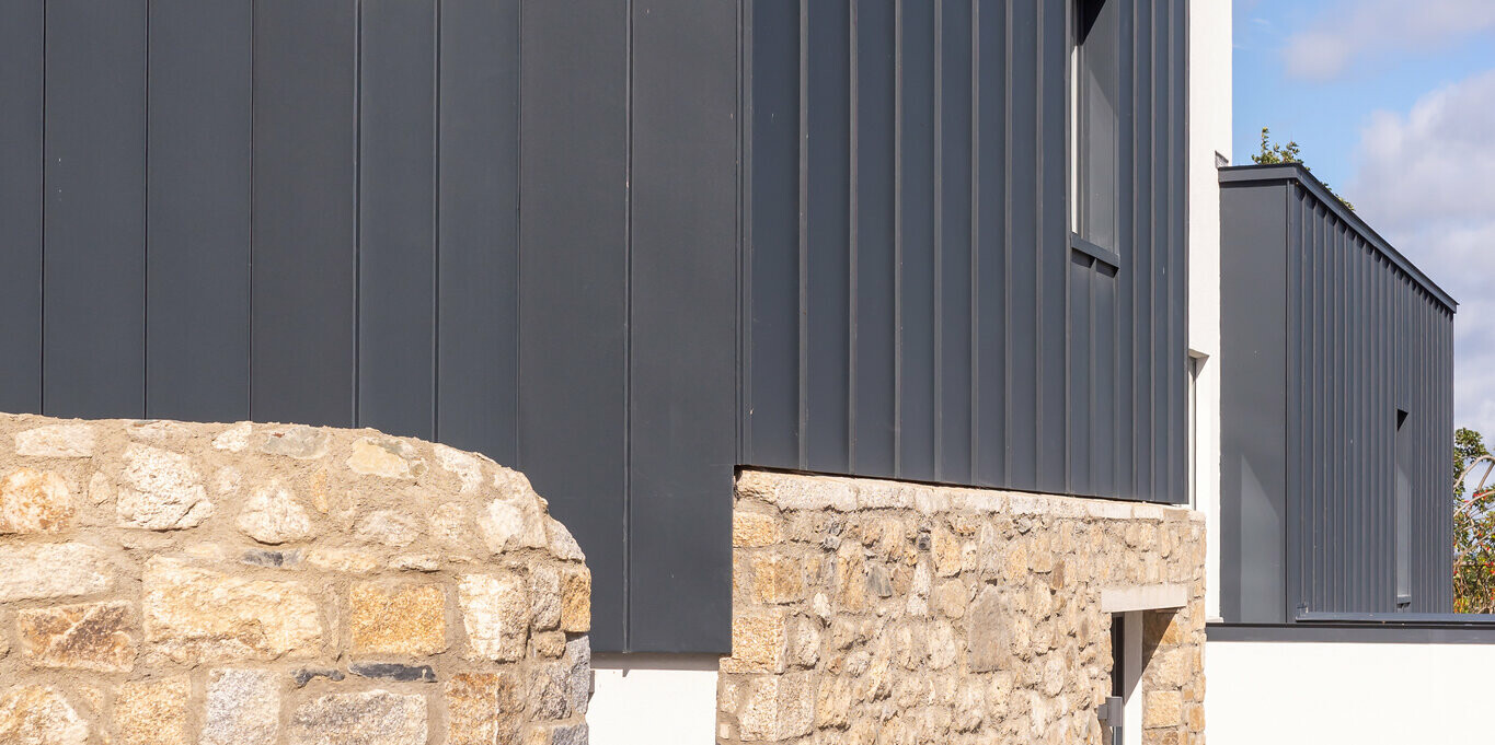Die Aufnahme zeigt die moderne Fassade eines Reihenhauses in Watson Place mit klaren Linien und einer markanten Kombination aus Natursteinmauer und dunkel anmutender PREFALZ Verkleidung in P.10 Anthrazit. Die stehenden Falze des Aluminiumprofils setzen vertikale Akzente und verleihen dem Gebäude eine elegante Struktur. Das witterungsbeständige PREFA Aluminium schützt dauerhaft vor Feuchtigkeit und Wind, während die leichte Bauweise flexible Gestaltung ermöglicht. Das Zusammenspiel aus robustem Naturstein und hochwertigem Aluminium schafft einen zugleich zeitlosen und dynamischen Gesamteindruck.