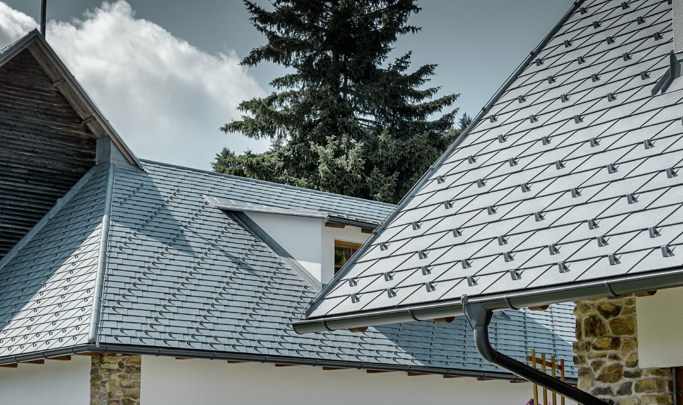 Detal aluminiowego krycia dachu PREFA, dachówka łupkowa w kolorze szarym kamiennym z aluminiową rynną dachową PREFA w kolorze antracytowym; w tle jest widoczna lukarna ciągniona z dachem na rąbek stojący. Elewacja jest biała z osadzonymi elementami kamiennymi.