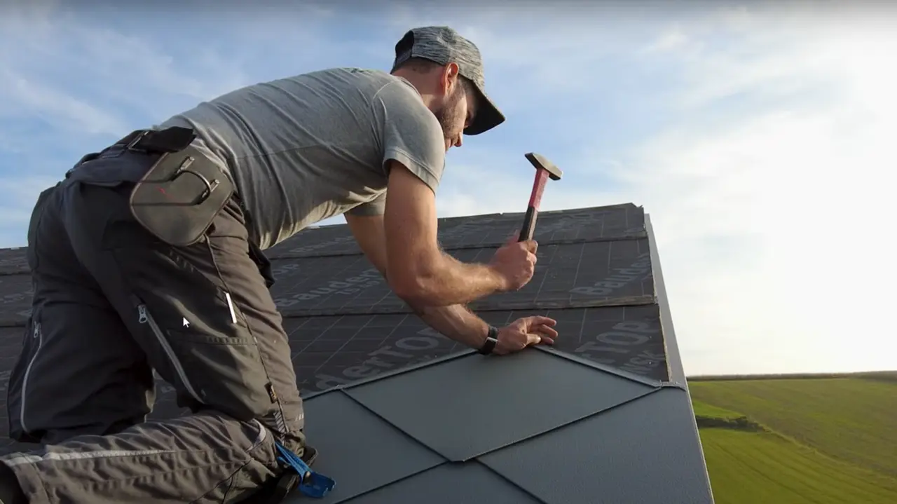 Blacharz w akcji: właściciel firmy „Der Blechmann” precyzyjnie układa dachówki PREFA romb 44 × 44 za pomocą młotka. Precyzję montażu widać na już ułożonych dachówkach na dolnej krawędzi zdjęcia. Mężczyzna ma na głowie czapkę i klęczy na dachu plecami do aparatu. W tle widać czyste niebo i zielony krajobraz. Harmonijne tło podkreśla symbolikę zdjęcia: trwałe i wytrzymałe rzemiosło z wykorzystaniem wysokiej jakości produktów aluminiowych PREFA.
