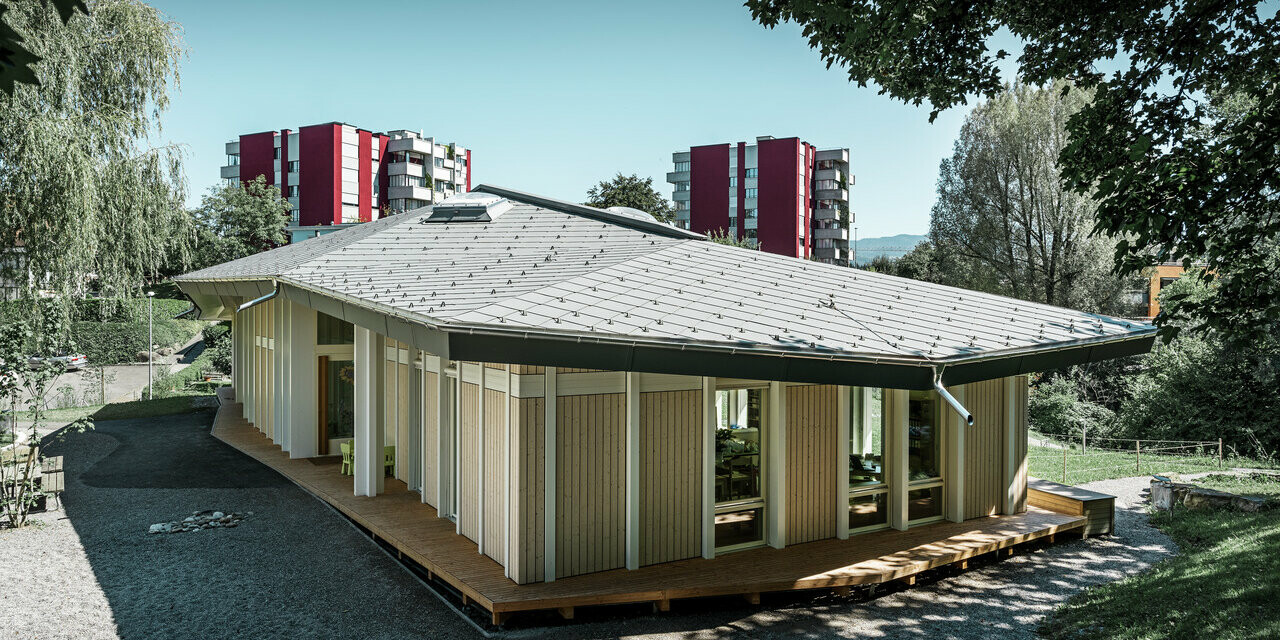 Jednokondygnacyjne przedszkole z pionowymi panelami elewacyjnymi i dachem aluminiowym PREFA (dachówka romb) w kolorze jasnoszarym