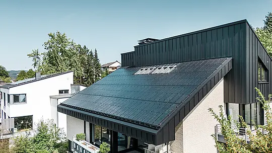 Nowoczesna architektura domu jednorodzinnego w Meschede w Niemczech, z dachem aluminiowym PREFALZ w kolorze P.10 czarny i zintegrowanymi modułami fotowoltaicznymi szkło/szkło. Dach z przesunięciem łączy w sobie ponadczasowy design z zrównoważoną technologią fotowoltaiczną. Innowacyjne moduły solarne PREFALZ zapewniają moc 18 kWp. Bezramowe moduły oraz eleganckie aluminium pokrycie dachowe i elewacyjne tworzą estetyczną i funkcjonalną całość, która harmonijnie wtapia się w zielone otoczenie.