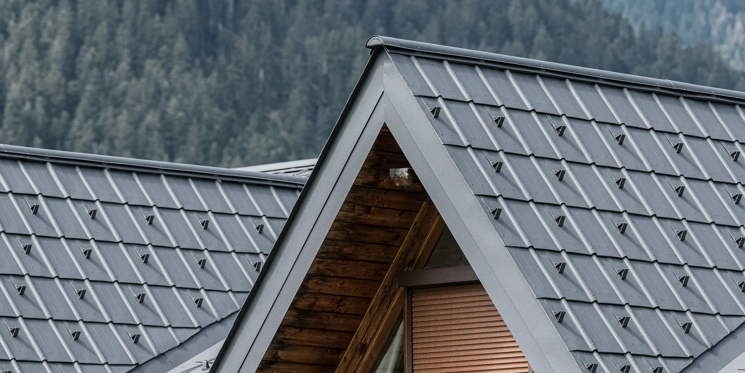 Nahaufnahme der Gaube auf dem Alpenhaus in Auronzo di Cadore, ausgestattet mit PREFA Ziegeln in Hellgrau. Die fein strukturierten Aluminiumpaneele bilden einen schönen Kontrast zum natürlichen Holz der Dachkonstruktion. Sie bieten einen hochwertigen Schutz vor dem alpinen Wetter und fügen sich nahtlos in die Bergwaldlandschaft ein.