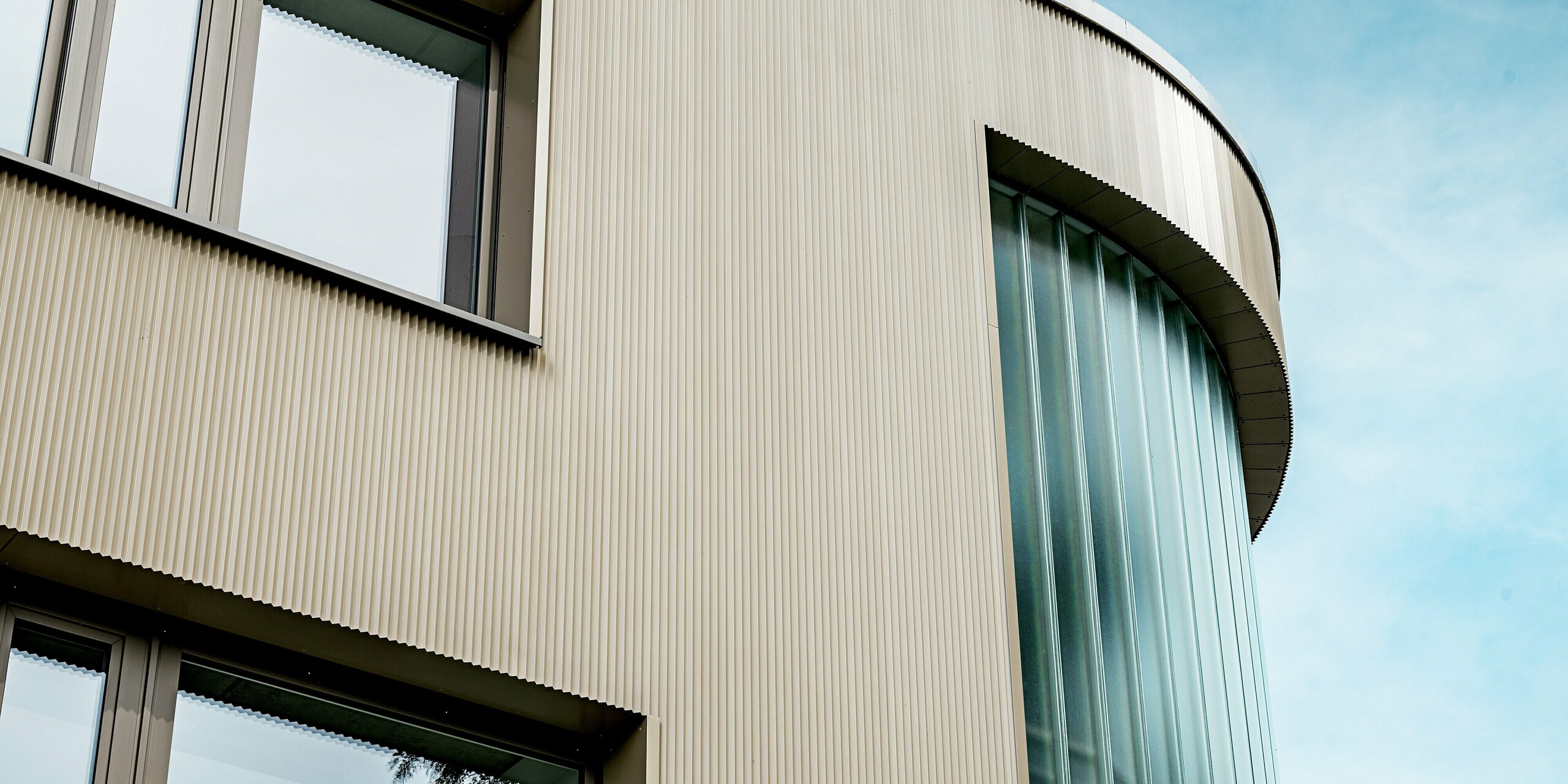 Das Bild zeigt einen faszinierenden Ausschnitt der Fassade des Festo-Gebäudes in Langenhagen, das mit PREFA Strangpressprofilen in Zackenform verkleidet ist. Die sanft geschwungene Gebäudekante und die vertikalen Glasflächen im Bereich der Ecke schaffen einen beeindruckenden Kontrast zur texturierten Aluminium-Fassade. Die klare Struktur der Zackenprofile in Bronze und das harmonische Zusammenspiel mit den großzügigen Fenstern und dem Glaselement verleihen dem Gebäude eine elegante, moderne Ausstrahlung. Diese Kombination aus funktionalen Materialien und ästhetischem Design sorgt für eine langlebige, pflegeleichte und gleichzeitig visuell ansprechende Architektur.