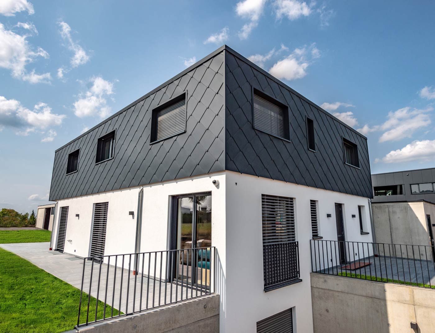 Rombowa płyta ścienna PREFA 44 x 44, antracytowa, referencja Nowoczesny nowy budynek z dachem płaskim — elewacja piętra została obłożona aluminiową rombową płytą ścienną PREFA 44 x 44 w kolorze antracytowym. Dolna część elewacji jest otynkowana w białym kolorze.